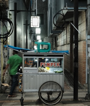 A small street food cart with a sign that reads 'PEMPEK' is situated in an alleyway with brick and concrete walls. The cart contains items for sale, and there is a chair on top. A blurred figure in a green shirt walks past, adding a sense of motion to the scene. Several illuminated signs hang above, providing light to the narrow passage.