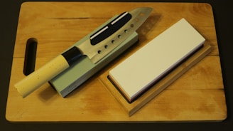 A kitchen knife with a beige handle rests on a sharpening stone, next to a whetstone on a wooden cutting board. The knife has multiple cutout holes in the blade.