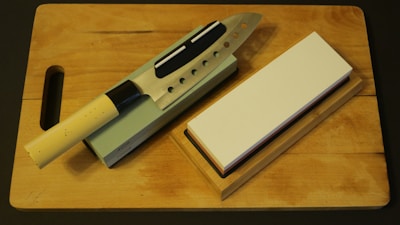 A kitchen knife with a beige handle rests on a sharpening stone, next to a whetstone on a wooden cutting board. The knife has multiple cutout holes in the blade.