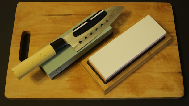 A kitchen knife with a beige handle rests on a sharpening stone, next to a whetstone on a wooden cutting board. The knife has multiple cutout holes in the blade.