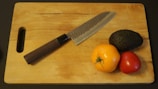 A wooden cutting board with a dark-handled knife lying diagonally across it. Next to the knife are three pieces of produce: a yellow tomato, a red tomato, and a dark green avocado.