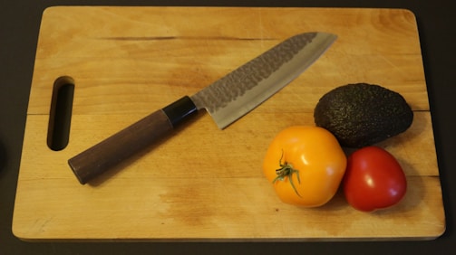 A wooden cutting board with a dark-handled knife lying diagonally across it. Next to the knife are three pieces of produce: a yellow tomato, a red tomato, and a dark green avocado.