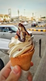A hand holding an ice cream cone with a swirl of vanilla and chocolate ice cream topped with chocolate syrup and a small chocolate piece with a logo. In the background, a busy street with cars and buildings.