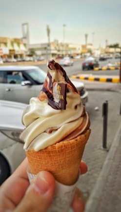 A hand holding an ice cream cone with a swirl of vanilla and chocolate ice cream topped with chocolate syrup and a small chocolate piece with a logo. In the background, a busy street with cars and buildings.