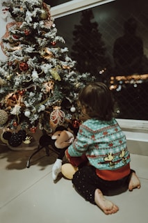 A cozy scene of a child wearing a festive Christmas sweater from mini and merry, smiling by a decorated tree.