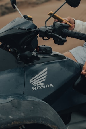 A close-up view of a Honda motorcycle's dashboard, showcasing the brand logo prominently on the body. A person holds a smartphone in their left hand, and the motorcycle's handlebars and controls are visible.
