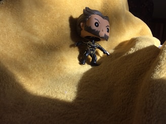 A small figure with cartoonish features and dark clothing stands on a textured yellow surface, possibly a blanket. The lighting creates pronounced shadows around the figure, emphasizing its features and the folds of the fabric.