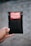 A hand is holding a small black bubble mailer against a blurred background. The mailer has a bright red and white label with the words 'HANDLE WITH CARE FRAGILE THANK YOU' printed on it.