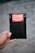 A hand is holding a small black bubble mailer against a blurred background. The mailer has a bright red and white label with the words 'HANDLE WITH CARE FRAGILE THANK YOU' printed on it.
