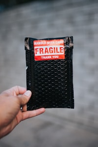 A hand is holding a small black bubble mailer against a blurred background. The mailer has a bright red and white label with the words 'HANDLE WITH CARE FRAGILE THANK YOU' printed on it.