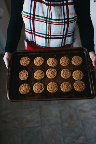 A person wearing a plaid apron is holding a baking tray filled with sixteen evenly spaced cookies. The cookies appear to be freshly baked and have a warm golden-brown color. They are placed on a dark, textured baking sheet in a kitchen setting with tiled flooring.