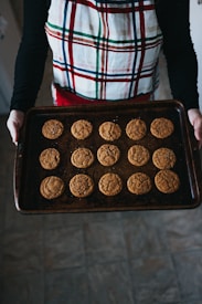 A person wearing a plaid apron is holding a baking tray filled with sixteen evenly spaced cookies. The cookies appear to be freshly baked and have a warm golden-brown color. They are placed on a dark, textured baking sheet in a kitchen setting with tiled flooring.