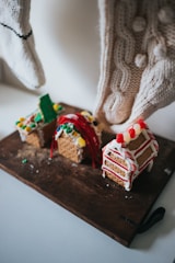 Three small gingerbread houses decorated with icing and candy are set on a wooden board. The houses are adorned with colorful candies, including red and white striped candy canes, and small gumdrops. The setup is cozy, with two cream-colored knit stockings hanging in the background.