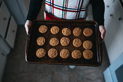 A person holds a baking tray filled with freshly baked cookies. The cookies appear to be soft and slightly cracked on the surface. The person is wearing a plaid apron and is in a kitchen with visible cabinets and floor tiles.