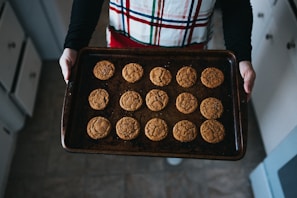A person holds a baking tray filled with freshly baked cookies. The cookies appear to be soft and slightly cracked on the surface. The person is wearing a plaid apron and is in a kitchen with visible cabinets and floor tiles.