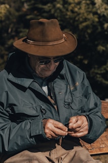 An elderly person is wearing a brown hat, sunglasses, and a heavy jacket, focusing intently on an activity, possibly tying something with their hands. They appear to be outdoors with a background of green foliage and a wooden bench.