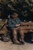 An older man wearing a brown hat and green jacket is seated on a wooden bench outdoors. He appears to be engaged in an activity, possibly examining or preparing a fishing line or lure. A fishing rod is propped next to him, and a camouflage pattern bag is on the ground beside the bench. The setting is natural with trees and greenery in the background.