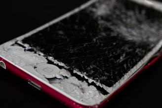A smartphone with a severely cracked and shattered screen, exposing the inner components. The body of the phone is red, and the damage suggests impact or pressure.