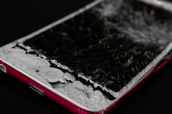 A smartphone with a severely cracked and shattered screen, exposing the inner components. The body of the phone is red, and the damage suggests impact or pressure.