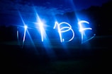Bright blue lights form the word 'VIBE' against a dark outdoor background, possibly captured using a long-exposure technique. The night sky and faintly visible trees blend into the scene.