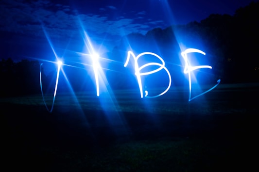 Bright blue lights form the word 'VIBE' against a dark outdoor background, possibly captured using a long-exposure technique. The night sky and faintly visible trees blend into the scene.