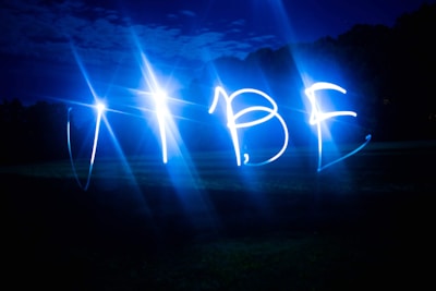 Bright blue lights form the word 'VIBE' against a dark outdoor background, possibly captured using a long-exposure technique. The night sky and faintly visible trees blend into the scene.