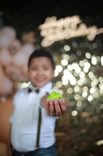 A child wearing a white shirt and suspenders holds a cupcake with bright green frosting toward the camera. The background is blurred with bokeh lights and pastel-colored balloons.