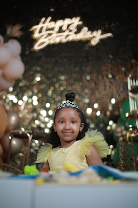 A young girl with a tiara on her head smiles while wearing a yellow dress with frilly shoulders. Birthday decorations are visible in the background, including balloons and a 'Happy Birthday' sign with illuminated lettering, set against a glittering backdrop.