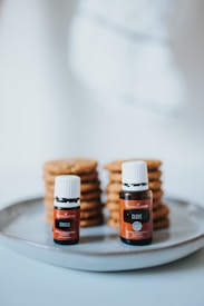 Two essential oil bottles, labeled Ginger and Clove, are placed on a gray plate. Behind the bottles, there are stacks of cookies or similar baked goods.