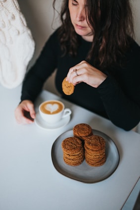 A person sits at a table holding a cookie above a plate with a stack of cookies. Next to the plate is a cup of cappuccino with a heart-shaped design in the foam. The person is wearing a black top and has medium-length, wavy brown hair.