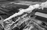 An aerial view of an industrial area with a large factory emitting dense white smoke from several chimneys. The factory is surrounded by multiple buildings, roads, and open land. There are also various vehicles and machinery visible around the site. The image is grayscale, adding a dramatic and somewhat somber mood.