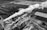An aerial view of an industrial area with a large factory emitting dense white smoke from several chimneys. The factory is surrounded by multiple buildings, roads, and open land. There are also various vehicles and machinery visible around the site. The image is grayscale, adding a dramatic and somewhat somber mood.