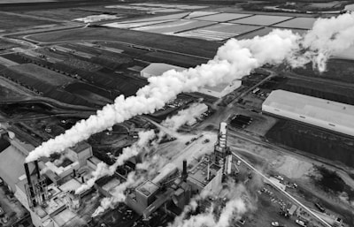 An aerial view of an industrial area with a large factory emitting dense white smoke from several chimneys. The factory is surrounded by multiple buildings, roads, and open land. There are also various vehicles and machinery visible around the site. The image is grayscale, adding a dramatic and somewhat somber mood.