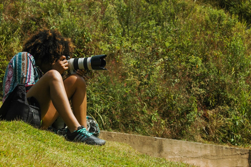 mulher em shorts pretos e tênis azuis sentados no banco de concreto segurando câmera dslr preta durante