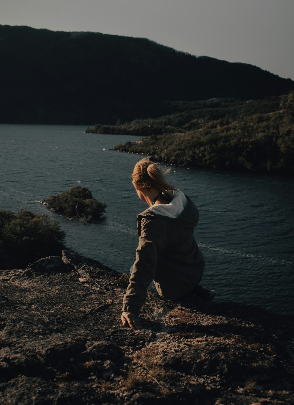 woman in gray jacket sitting on rock near body of water during daytime