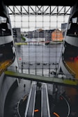 A spacious interior with modern architecture featuring large escalators and a bridge walkway. The front glass wall offers a view of a river and urban buildings outside. The space has a sleek design with concrete and glass elements.