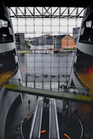 A spacious interior with modern architecture featuring large escalators and a bridge walkway. The front glass wall offers a view of a river and urban buildings outside. The space has a sleek design with concrete and glass elements.