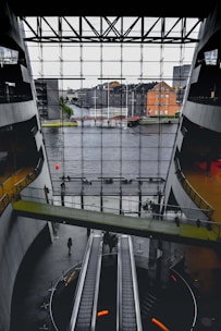 A spacious interior with modern architecture featuring large escalators and a bridge walkway. The front glass wall offers a view of a river and urban buildings outside. The space has a sleek design with concrete and glass elements.