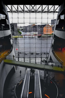 A spacious interior with modern architecture featuring large escalators and a bridge walkway. The front glass wall offers a view of a river and urban buildings outside. The space has a sleek design with concrete and glass elements.