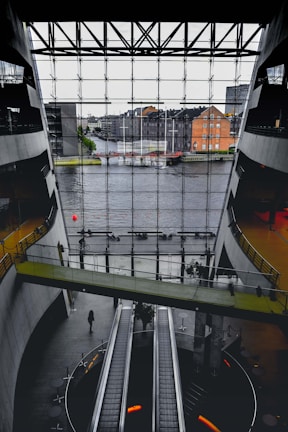 A spacious interior with modern architecture featuring large escalators and a bridge walkway. The front glass wall offers a view of a river and urban buildings outside. The space has a sleek design with concrete and glass elements.
