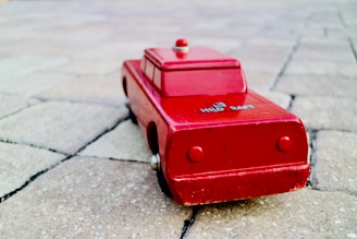 A vintage red toy car made of wood with simple, rounded features. It is positioned at an angle on a textured stone surface, casting a soft shadow. The toy is slightly worn, showing signs of age and play.