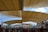 A large crowd of people gathers under an expansive, modern architectural structure featuring a series of tan, triangular fabric canopies. The structure creates a shaded walkway, and the bright blue sky with scattered clouds is visible above. Some flags and architectural supports are visible along the sides.