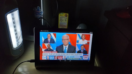 A tablet screen displays a CNN news segment showing results for the 2020 U.S. presidential election with Joe Biden elected president. The broadcast features six separate images of people, including a central anchor and participants in smaller screens. The headline reads 'BREAKING NEWS' and discusses Trump's lack of public acceptance. The setup includes a lit white lamp on the left and a box of crackers sitting behind the tablet.