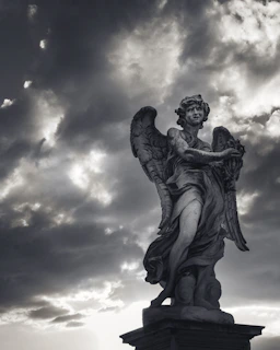 A stone statue of an angel is prominently displayed against a dramatic sky filled with dark, swirling clouds. The angel's wings are intricately carved, and its expression is solemn, adding a sense of majesty to the scene.