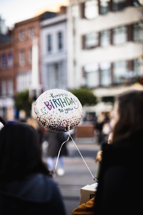 A balloon with the phrase 'happy birthday to you' floats in a street setting. The balloon is colorful with multicolored dots. Blurred people and urban buildings are visible in the background.