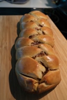 A golden-brown braided bread loaf with a glossy finish rests on a wooden cutting board. The bread appears freshly baked, with the braided sections revealing a rich, brown filling.