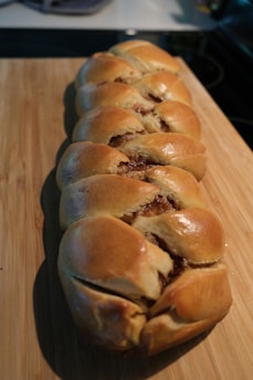 A golden-brown braided bread loaf with a glossy finish rests on a wooden cutting board. The bread appears freshly baked, with the braided sections revealing a rich, brown filling.