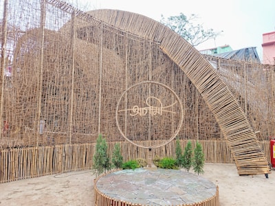 An intricate bamboo structure with a large archway and woven design. A circular frame with a stylized inscription is placed prominently at the center. Small green plants are arranged near the base, and the setting appears to be part of an outdoor area.
