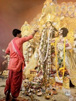 A man dressed in traditional clothing is performing a ritual in front of an elaborately decorated idol adorned with garlands and intricate ornaments. The idol is part of a larger structure with detailed and ornate designs, rich in gold color. The floor is covered with various offerings, including flowers and small containers.