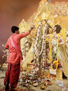 A man dressed in traditional clothing is performing a ritual in front of an elaborately decorated idol adorned with garlands and intricate ornaments. The idol is part of a larger structure with detailed and ornate designs, rich in gold color. The floor is covered with various offerings, including flowers and small containers.